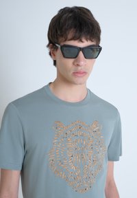 Camiseta de color azul claro hecha de algodón con un diseño de león cortado con láser en color beige. El modelo lleva gafas de sol rectangular oscuras y tiene el cabello corto.
