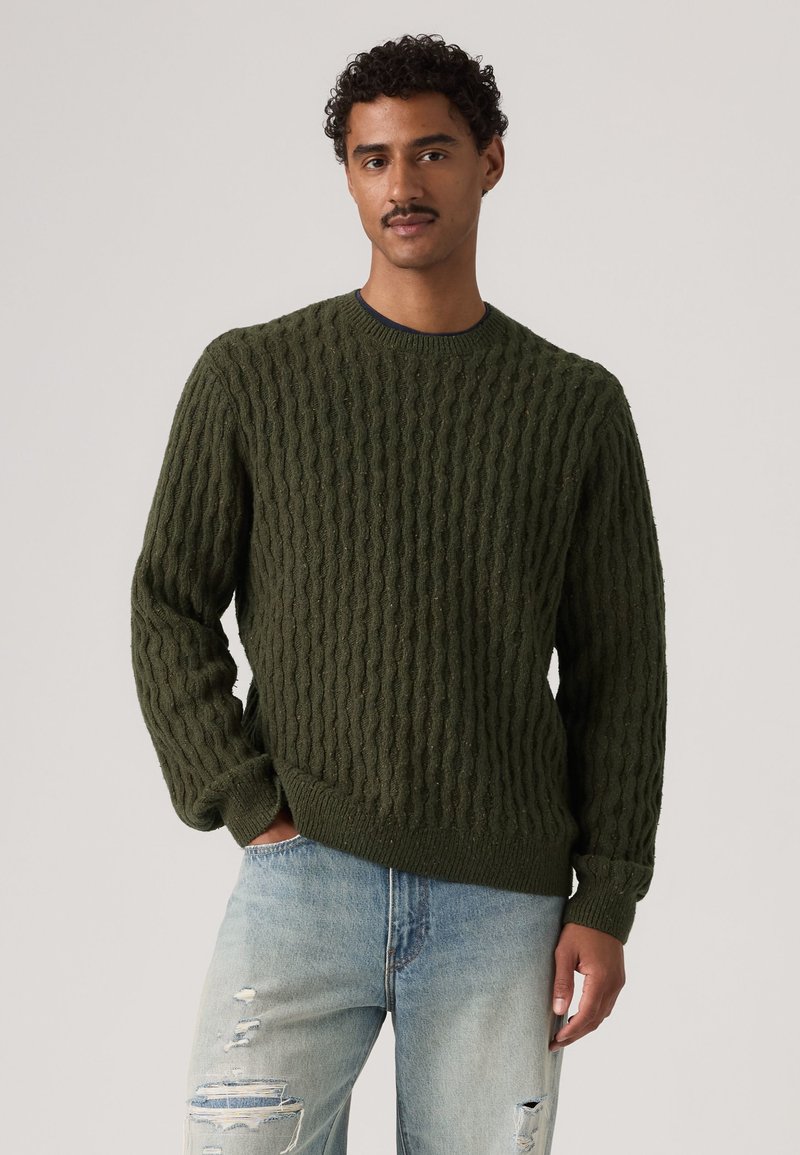 Homme aux cheveux bouclés et moustache porte un pull vert en maille torsadée et un jean bleu clair déchiré, debout avec une main dans la poche.