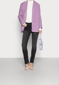 Kvinna klädd i en lila kavaj, vit polotröja, svarta skinny jeans, vita öppna klackskor, som håller en liten lavendelfärgad handväska vid sin sida.