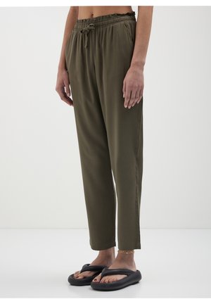 Persoon draagt olijfgroene losse broek met elastische tailleband en zwarte gevoerde teenslippers, staand tegen een effen witte achtergrond.