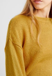 Pletený hořčicově žlutý svetr s volným střihem, žebrovaným výstřihem a strukturovanou tkaninou. Viditelné stehy vytvářejí jemný, útulný vzhled.