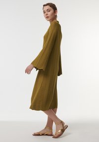 Robe vert olive avec texture en vagues, manches cloches longues, ourlet mi-long. Sandales plates dorées avec fines lanières. Design simple et élégant.