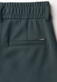 Tissu extensible vert foncé avec une taille élastique froncée. Présente une seule poche arrière et un accent logo en métal. Texture lisse.