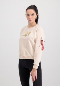 Béžová mikina s okrúhlym výstrihom, zlatým logom "Alpha Industries" a utility vreckom s červenou nášivkou na ľavom rukáve; mäkká textúra, voľný strih.