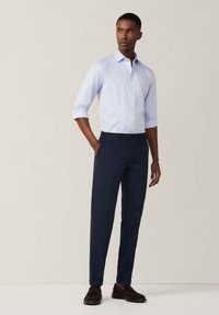 Camisa de botones de manga corta en azul claro, con cuello, combinada con pantalones de traje en azul marino oscuro y zapatos marrones, de pie sobre un fondo neutro.