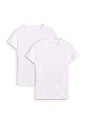 Młoda kobieta w białym t-shircie, jasnoszarych dresach, szarych trampkach i czarnej czapce, stoi z jedną ręką w kieszeni.