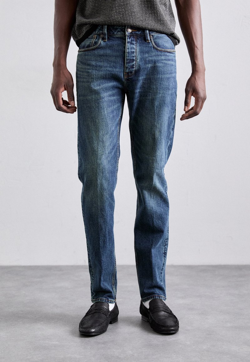 Jeans in denim blu con gamba dritta, lavaggio chiaro e sbiaditure sottili, abbinati a mocassini neri intrecciati e un top grigio a fantasia.
