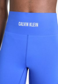 Gros plan sur une personne portant des leggings taille haute bleu vif avec le logo "Calvin Klein" imprimé en blanc sur la ceinture.