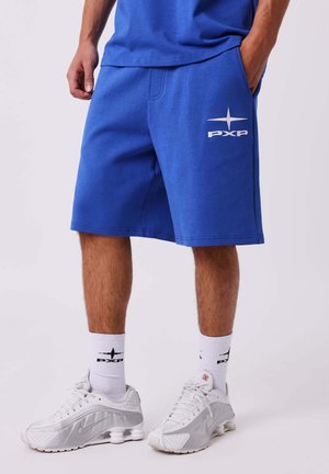 Man draagt blauwe PXP-shorts, witte PXP-sokken en witte Nike-sneakers, staande met één hand in de zak tegen een witte achtergrond.