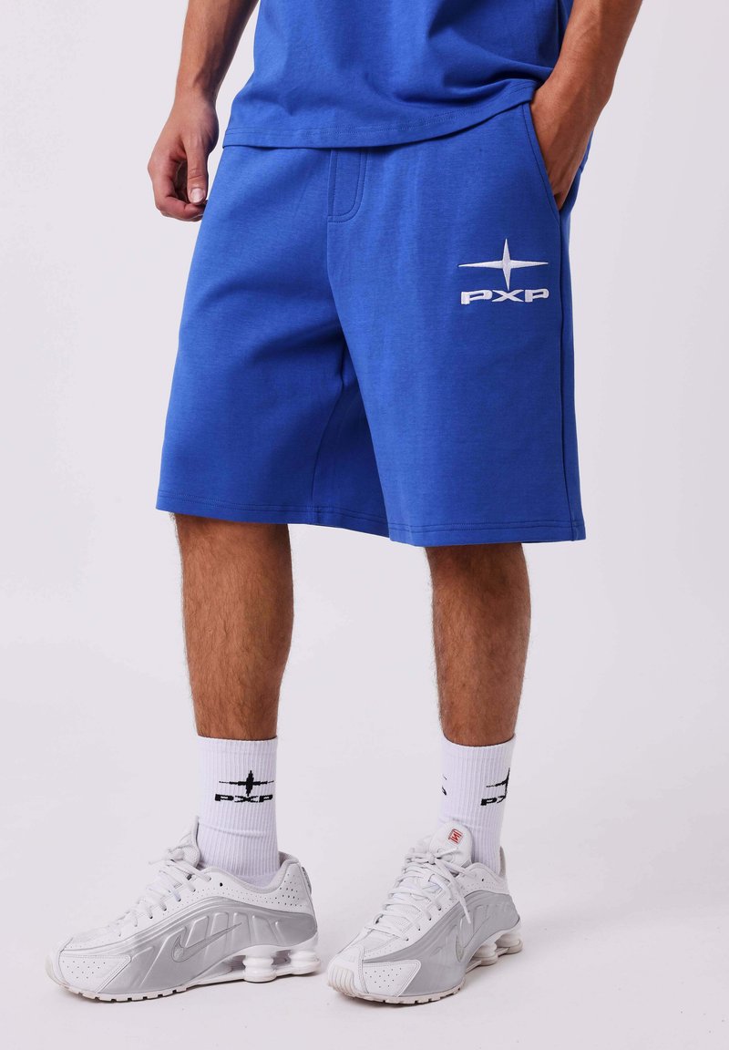 Homme portant un short bleu PXP, des chaussettes blanches PXP et des baskets blanches Nike, debout avec une main dans la poche sur fond blanc.