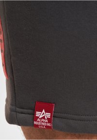 Tissu de vêtement foncé, montrant une étiquette rouge avec le logo blanc Alpha Industries Inc U.S.A. et du texte près de l'ourlet.