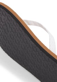 Tongs avec une lanière en tissu blanc et une semelle en caoutchouc noir texturé avec un bord marron, mettant en valeur un motif en nid d'abeille.