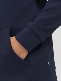 Le sweat-shirt bleu marine présente une texture douce, des poignets côtelés et une poche avant avec une petite étiquette de logo sur le côté.