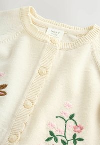 Helekreemjas kampsun, millel on roosades ja pruunides toonides lilleline tikand, ümar kaelus ning eest nööbitav sulgemine laineliste ääre detailidega.