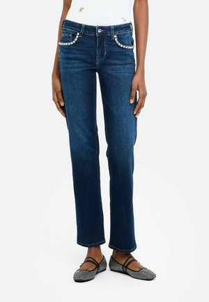 Dark blue straight-leg jeans with rhinestone detailing on front pockets worn with studded black flat shoes.