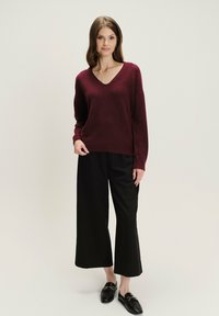 Vrouw in een bordeauxrode V-hals trui, zwarte wijde cropped broek en zwarte instappers, staande tegen een effen lichte achtergrond.