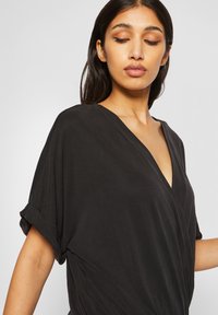 Femme aux longs cheveux foncés portant un chemisier noir ample avec des manches retroussées, regardant vers le bas, les yeux fermés.