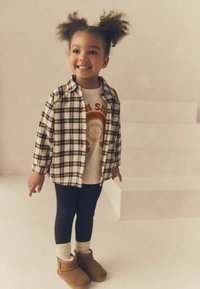 Vaikas vilki kvadratinį flanelinį marškinį kreminės, rudos ir tamsiai mėlynos spalvų, užsidėjęs grafišką marškinėlį, juodas leginsus ir rudus ankle batus su minkštu paviršiumi.