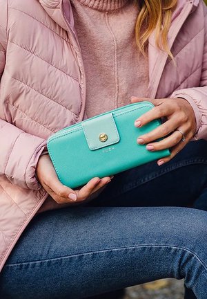 Turquoise wallet made of textured material, featuring a snap closure with a gold button, held in a hand against a pink jacket and dark jeans.