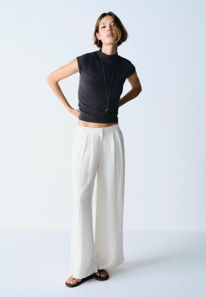 Fiatal nő áll, kezét a csípőjén tartva, fekete ujjatlan felsőt, hosszú nyakláncot, bő fehér nadrágot és fekete szandált viselve, fehér háttér előtt.