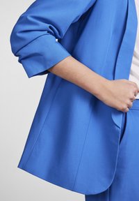 Blazer blu su misura con texture liscia, maniche ripiegabili e silhouette pulita, dotato di tasche laterali e taglio moderno.