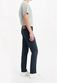 Mörkblå jeans med rak skärning och en beige läderlapp vid midjan; kombinerat med en ljusgrå t-shirt med kort ärm och vita sneakers.