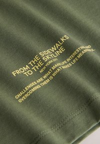 Maslinasto zelena majica s teksturiranim materijalom, žutim printanim tekstom i detaljima šavova. Tekst prenosi poruke o izazovima i gradskom životu.