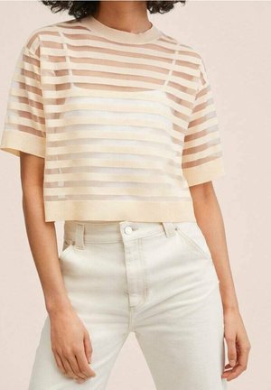 Kvinde iført en gennemsigtig, kortærmet crop top med beige og hvide horisontale striber over en hvid camisole og lysebeige højtaljede bukser.