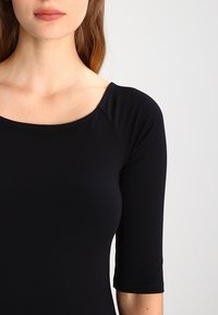 Svart välsittande topp med båtringning och trekvartslånga ärmar. Tillverkad av ett slätt, elastiskt material, har den en sömlös design.