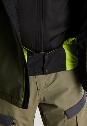 Nærbillede af person iført sort jakke med neongrøn inderforing og beige bukser med knappeklap og lynlåsdetaljer.