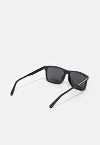 Occhiali da sole neri con lenti rettangolari, montatura opaca e tempie sottili. Presentano un design minimalista con lenti grigie oscurate.