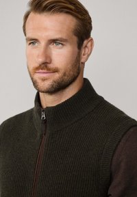 Olivově zelená pletená vesta s vysokým límcem a předním zipem. Žebrovaná textura a kontrastní hnědý zip obohacují design.