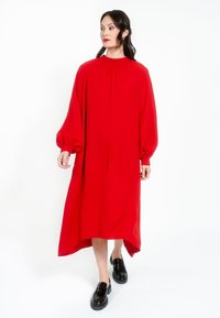 Červené šaty s dlouhým rukávem, vysokým límcem a nařasením, s uvolněnou siluetou a asymetrickým lemem, doplněné černými botami.