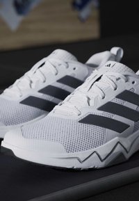 Ζευγάρι λευκά παπούτσια τρεξίματος Adidas με μαύρες ρίγες, επάνω μέρος από δίχτυ και σόλα με ζιγκ-ζαγκ σε σκοτεινή επιφάνεια.