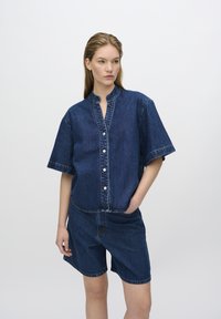 L'outfit in denim presenta una camicia blu con bottoni e maniche corte, abbinata a un paio di pantaloni corti fino al ginocchio. Dettagli di cucitura visibili.