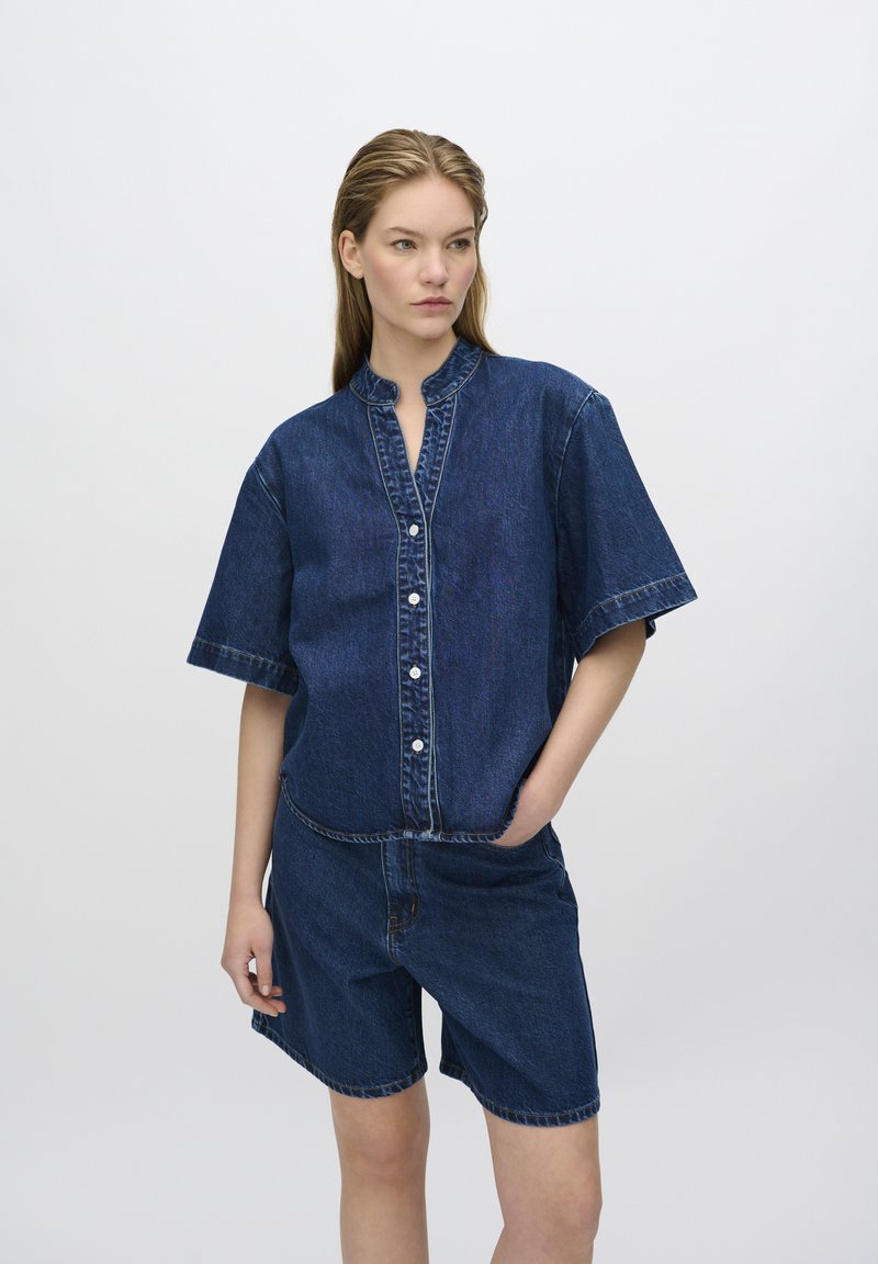 L'outfit in denim presenta una camicia blu con bottoni e maniche corte, abbinata a un paio di pantaloni corti fino al ginocchio. Dettagli di cucitura visibili.
