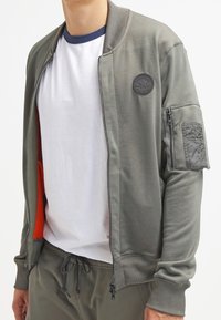 Giubbotto bomber verde oliva con polsini e colletto a costine, chiusura con zip sul davanti e una patch sulla manica sinistra. L'interno è foderato in arancione.