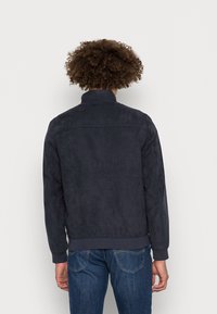 Granatowa zamszowa kurtka z kołnierzem typu stójka, ściągaczami przy rękawach i na dole. Gładka faktura i minimalistyczny design. Noszona na dżinsach.