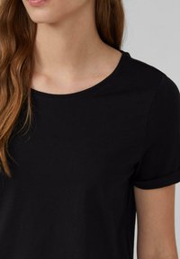 T-shirt noir avec un col rond et des manches courtes repliées, fabriqué en tissu de coton doux avec une texture lisse. Les détails de couture sont visibles.