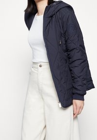 Granatowa pikowana kurtka z kapturem, z elastycznymi mankietami i naszywką z logo. Nosi się ją na biały top w prążki i kremowe spodnie.