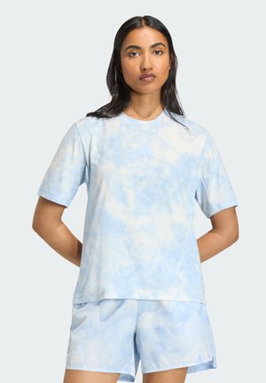 Frau trägt hellblaues, batikgefärbtes T-Shirt mit kurzen Ärmeln und passende Shorts, steht mit den Händen hinter dem Rücken vor einfarbigem Hintergrund.