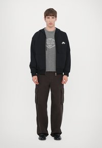 Felpa nera con zip sopra una felpa grigia con grafica, abbinata a pantaloni cargo marroni con tasche laterali, completata da scarpe nere.
