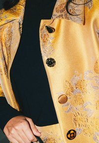 Giacca in satin con motivo floreale dorato e top nero sotto. Presenta bottoni rotondi ambrati e un caratteristico bottone a forma di fiore nero.