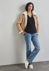Giacca di camoscio marrone chiaro con colletto in shearling bianco, top nero a scollo a V, jeans blu e sneaker bianche, caratterizzata da una vestibilità comoda e una texture liscia.