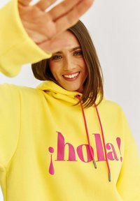 Žltá mikina s kapucňou, na ktorej sa nachádza výrazný ružový nápis "¡hola!" s akcentom. Navrhnutá so voľným strihom a sťahovacou šnúrkou. Hladká textúra.