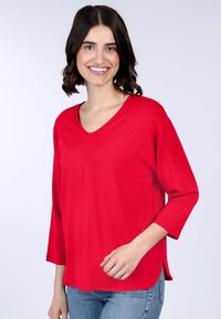 Rotes V-Ausschnitt-Oberteil mit 3/4-Ärmeln, aus glattem Stoff gefertigt. Verfügt über einen entspannten Schnitt und seitliche Schlitze für zusätzlichen Komfort. Getragen mit blauen Jeans.