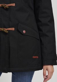 Schwarze Außenjacke aus strukturiertem Stoff mit hölzernen Knebelknöpfen und einer seitlichen Tasche mit einem markierten braunen Lederanhänger.