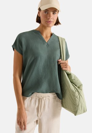 Mladá žena s béžovou čepicí, zeleným krátkým rukávem a světle béžovými kalhotami, držící přes rameno velkou prošívanou světle zelenou tašku.