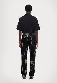 Melnas denima bikses ar balināšanas pleķu rakstu, ar standarta aizmugurējām kabatām un melnu ādas uzlīmi jostasvietā. Struktūrveidaaudums.