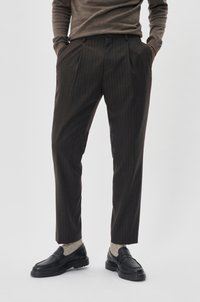 Braune Nadelstreifenhose mit taillierter Passform, versehen mit Falten an der Vorderseite. Kombiniert mit dunklen Slippern und hellen Socken.